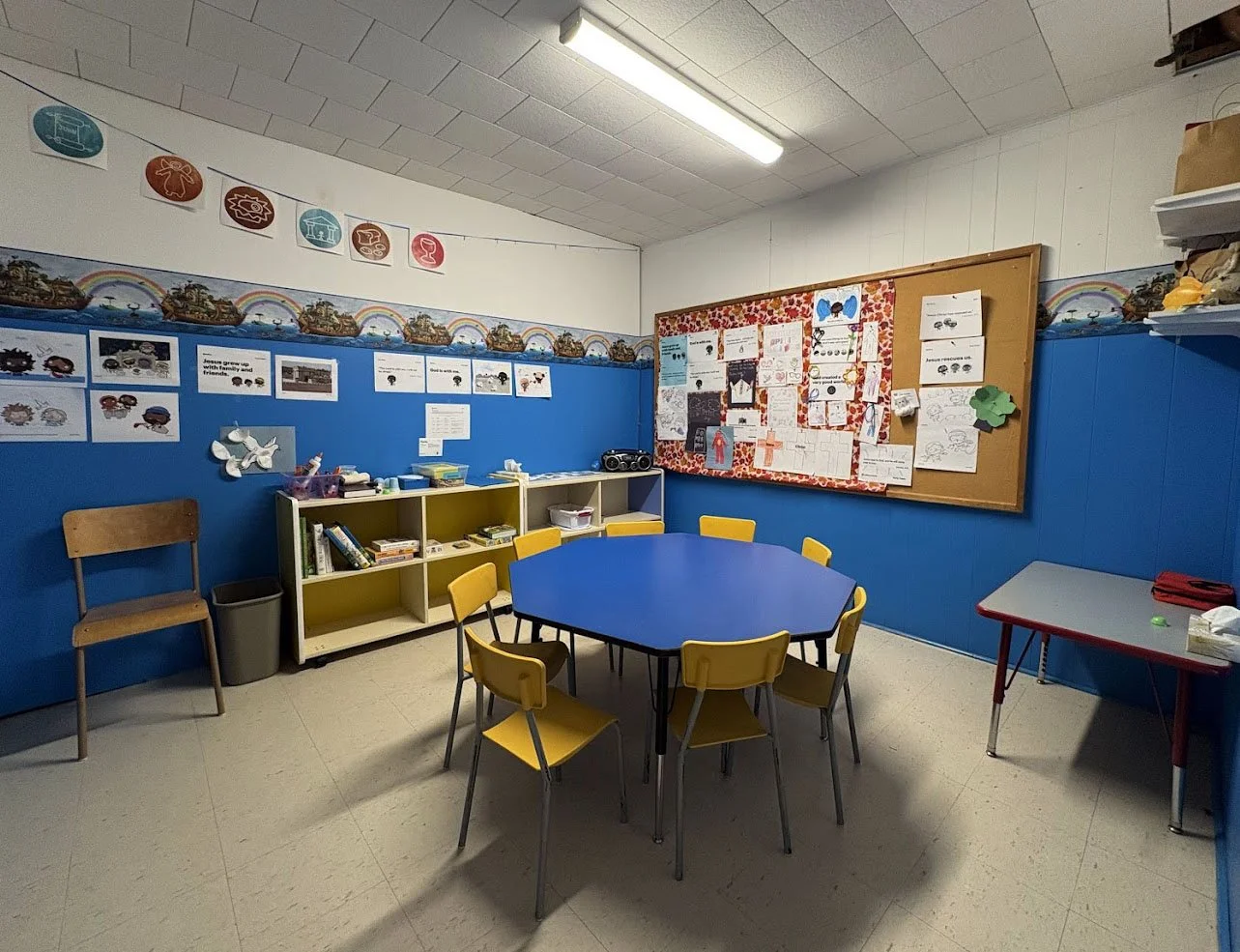 Classroom with a blue hexagonal table surrounded by six yellow chairs, a bulletin board with children's artwork, and shelves with books and supplies, all in a room with white walls and a blue half-wall.