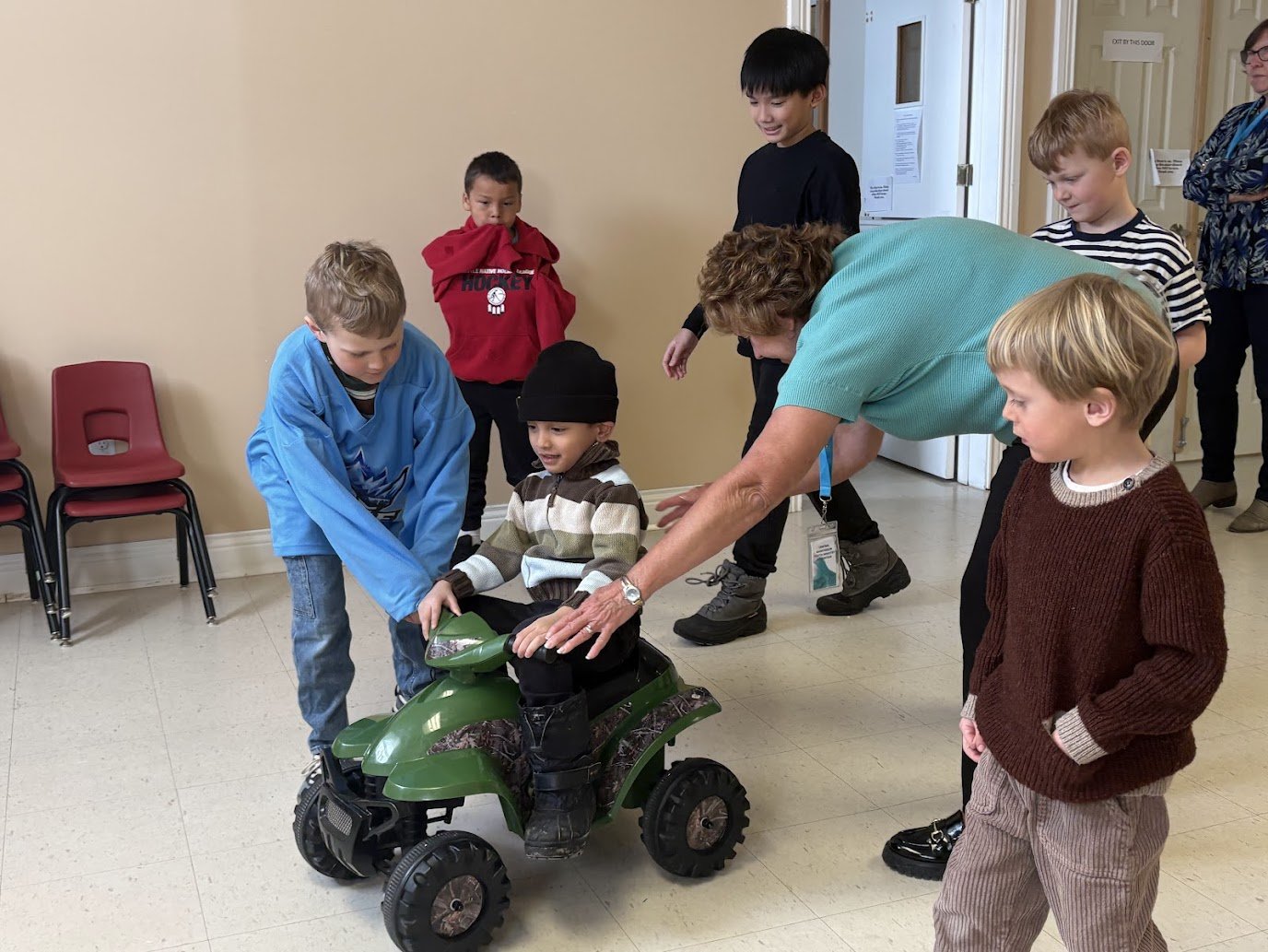Young boy sitting on a small green toy quad bike, with a woman assisting him as other children watch nearby.