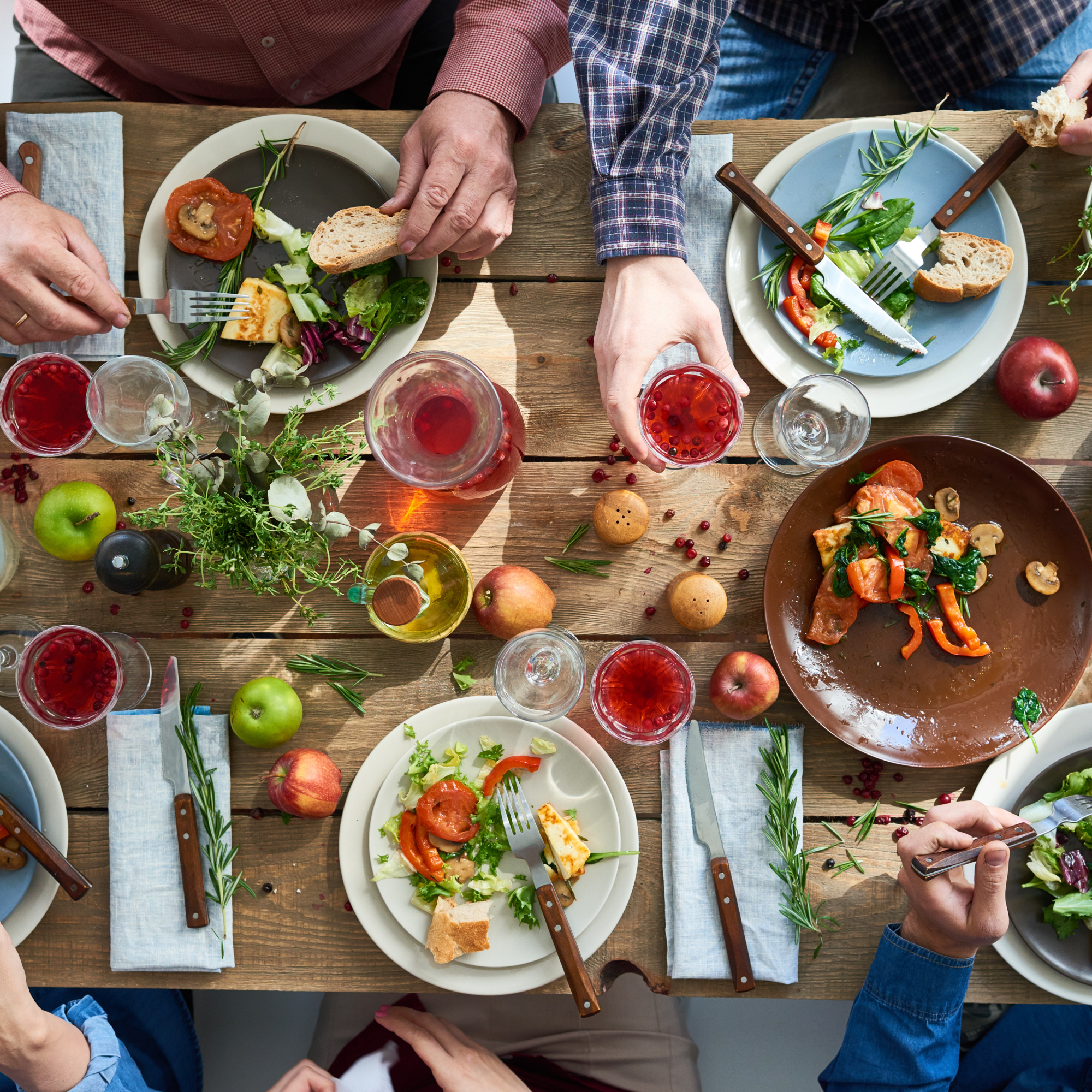 A top-down view of a wooden dining table with various plates of salad, bread, apples, and glasses of red drinks, surrounded by people reaching for food and drinks.