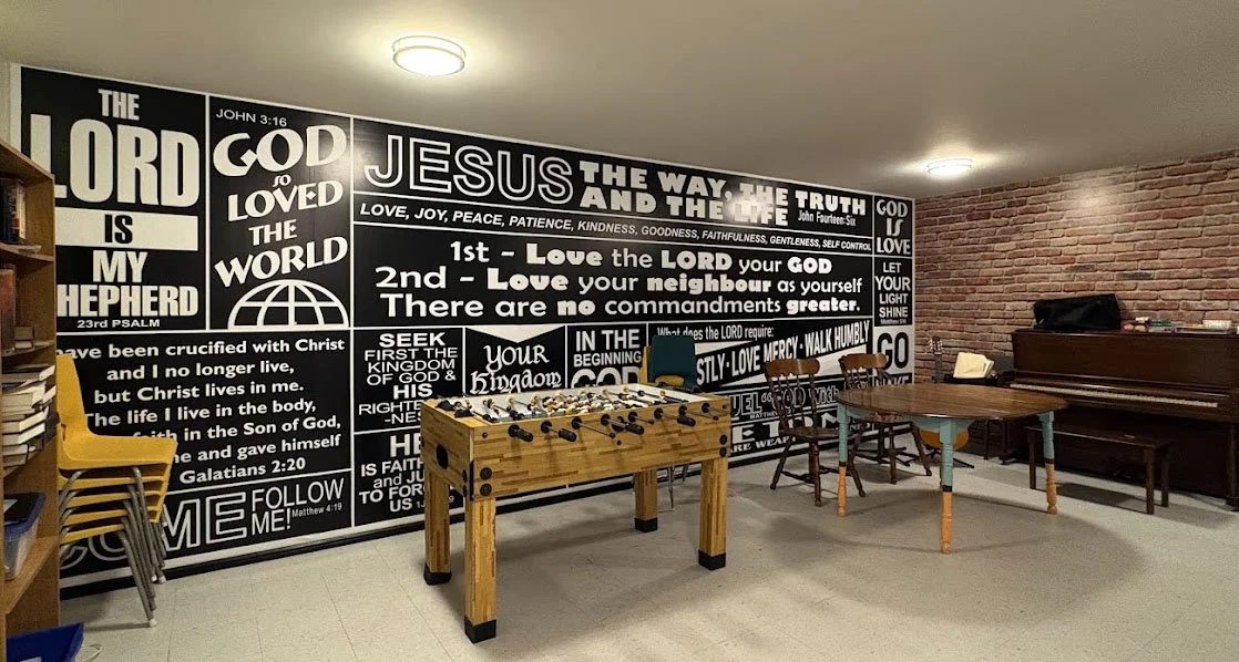 Indoor room with a large black-and-white wall mural featuring Bible verses and Christian messages. There is a foosball table, wooden chairs, and a piano.