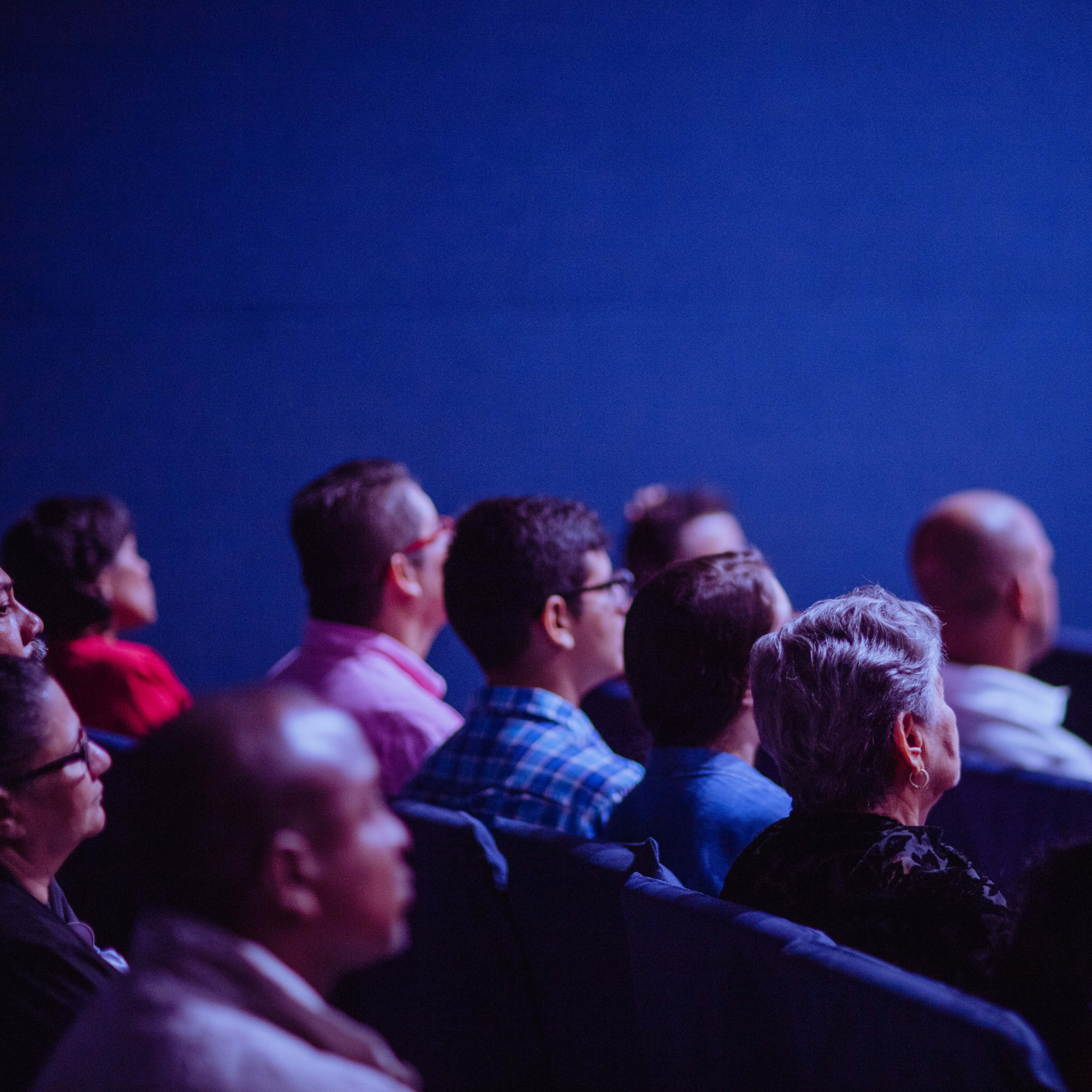 Audience of diverse adults seated in a dark theater or conference room watching a presentation.