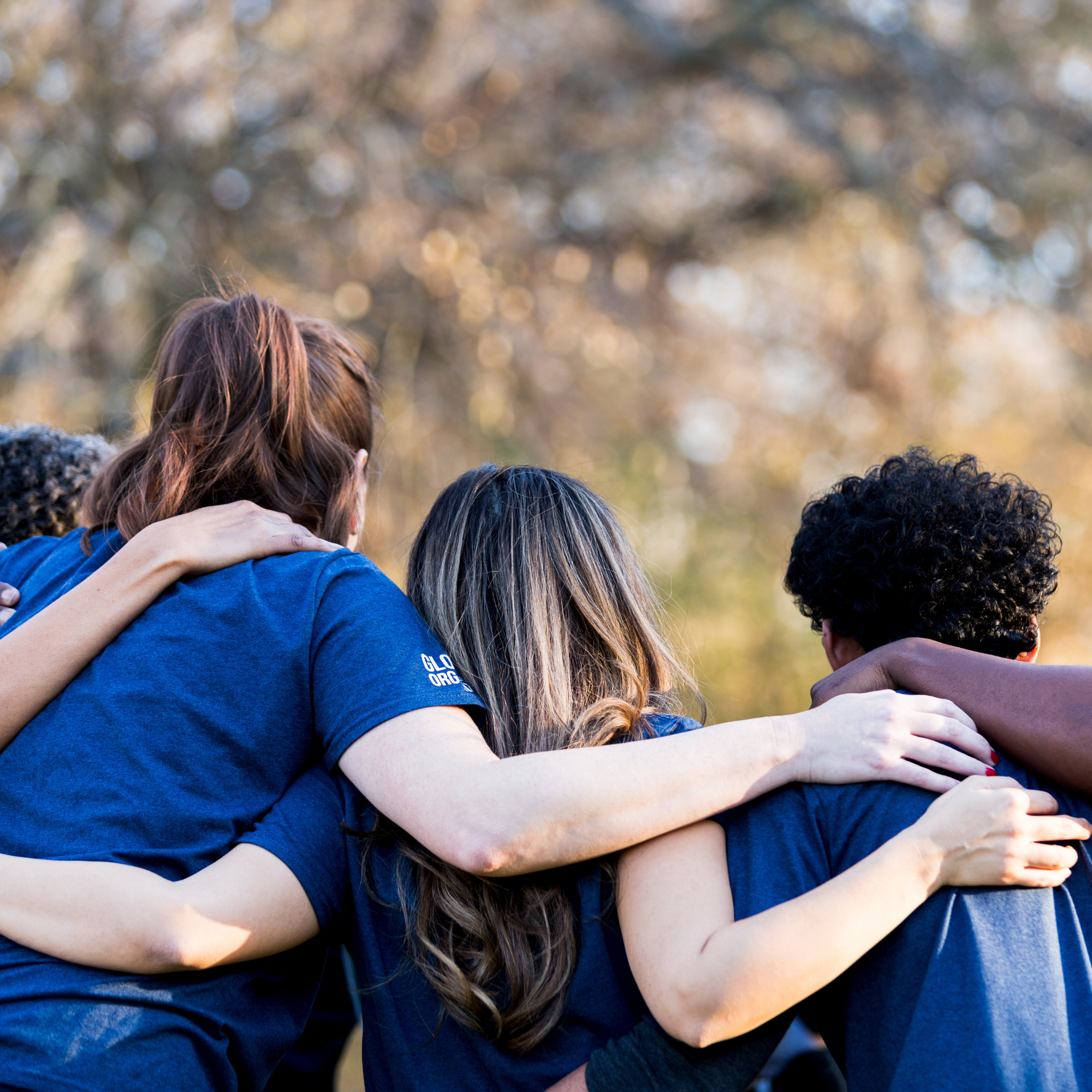 Group of diverse women and girls with arms around each other, huddled and standing outdoors.