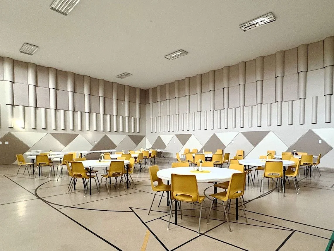 Empty large indoor room with several round tables and yellow chairs, gymnasium type flooring, and geometric wall patterns.