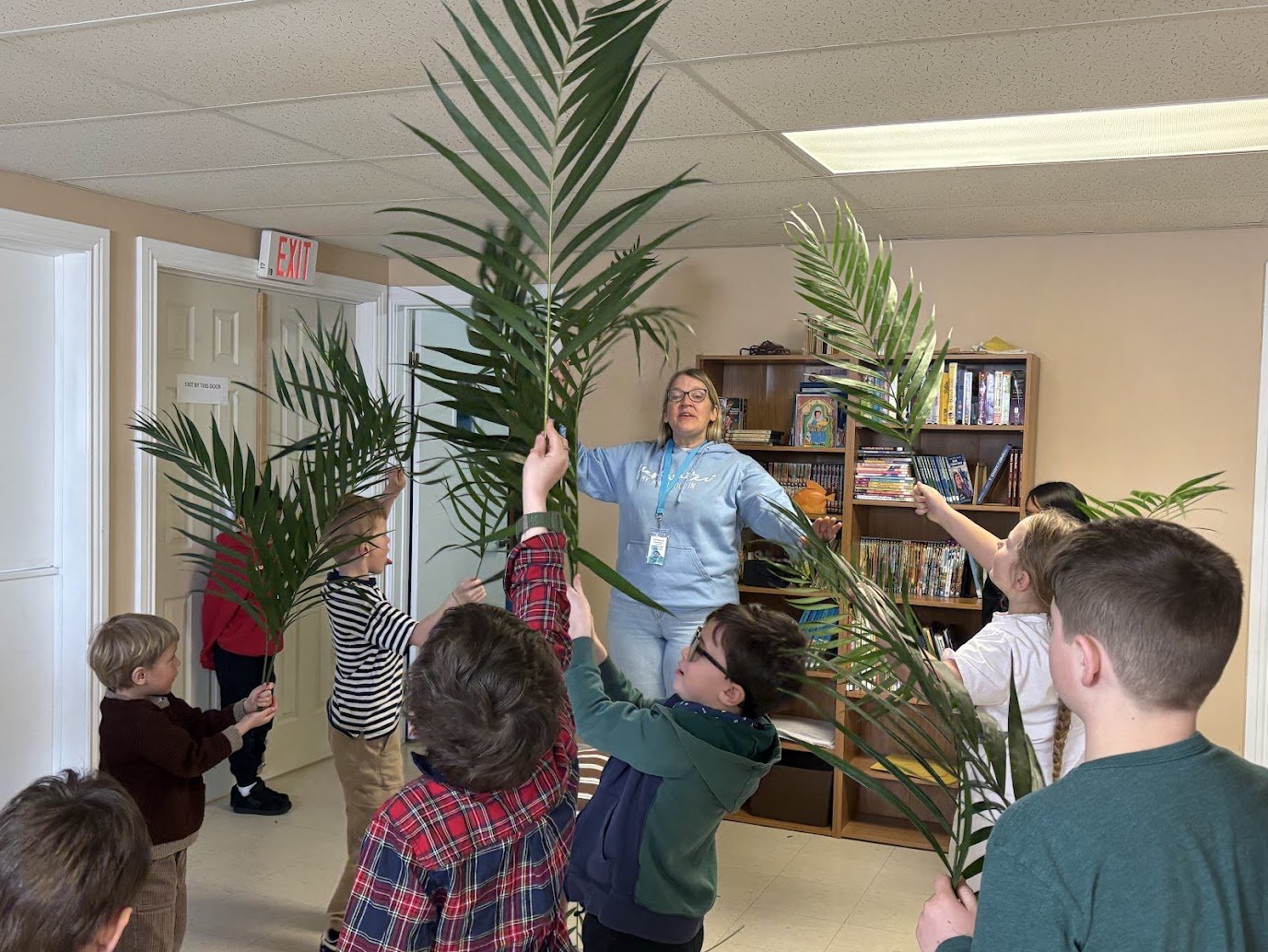 Group of children holding large palm leaves around a woman in a classroom or library setting.