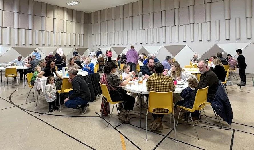 People sitting at round tables in a gymnasium or community center, eating and talking during a group event or gathering.