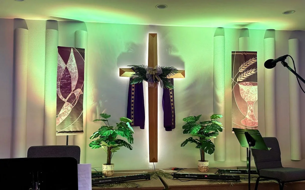 Church altar with a wooden cross decorated with greenery and a bow, flanked by two potted plants, illuminated with green lighting, with banners featuring chalices and wheat on either side, and microphones and chairs in the foreground.
