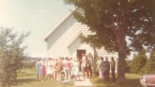 A group of people gathered outside a white church with a steep roof, under a large tree, during the daytime.