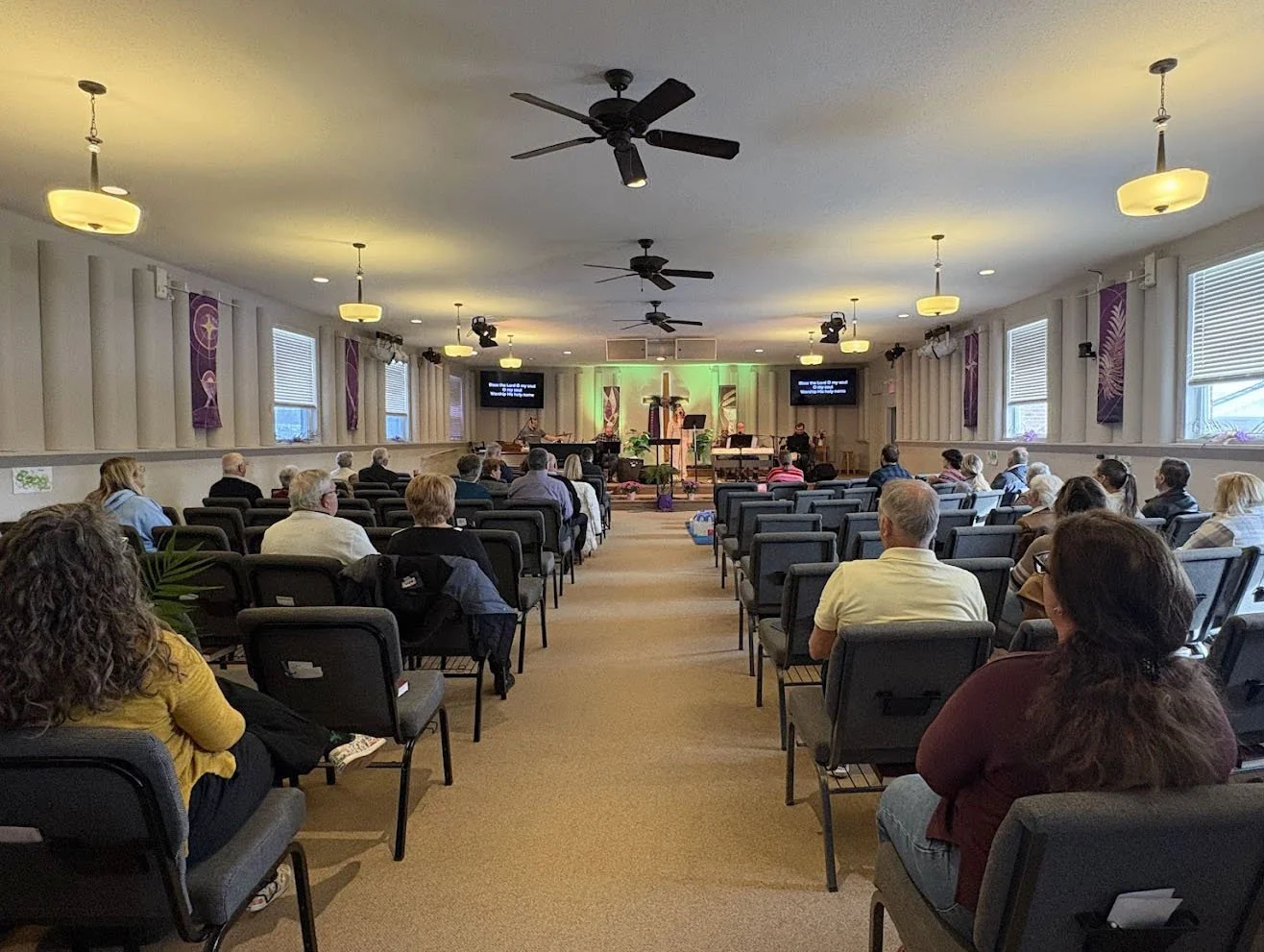 Inside a church with people seated facing the altar where a speaker is presenting. The room is lit by ceiling lights and has windows on both sides.