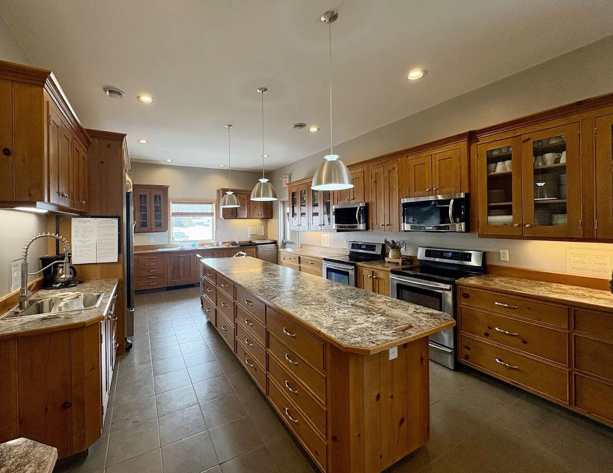 A spacious kitchen with wooden cabinets, granite countertops, stainless steel appliances including a microwave, oven, and refrigerator, three pendant lights above a kitchen island, and a window above the sink