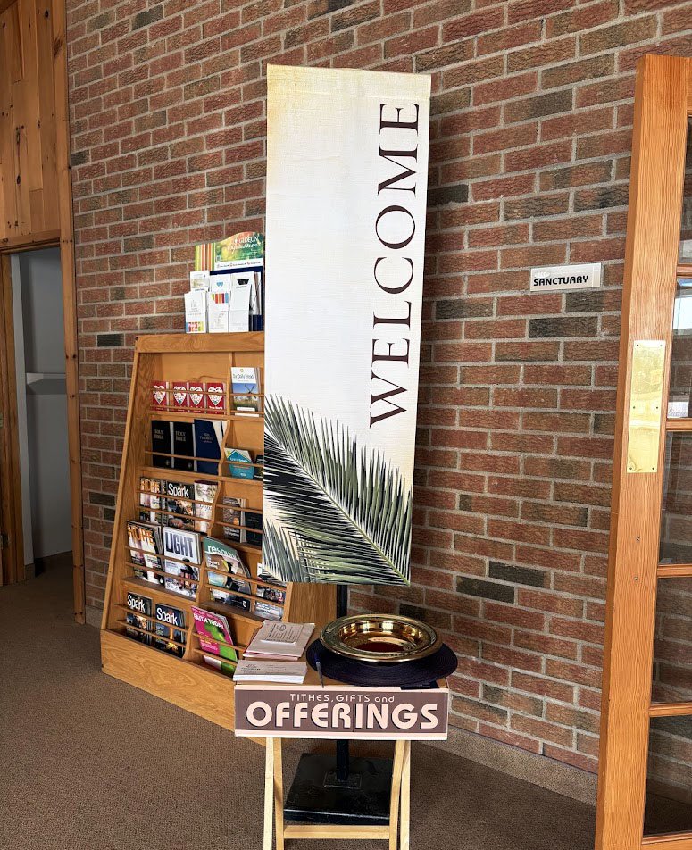 A welcome sign with a palm leaf design leaning against a brick wall, next to a display rack with religious booklets, and a small table with a tray, a sign that says "Tithes, Gifts and Offerings," and some papers.