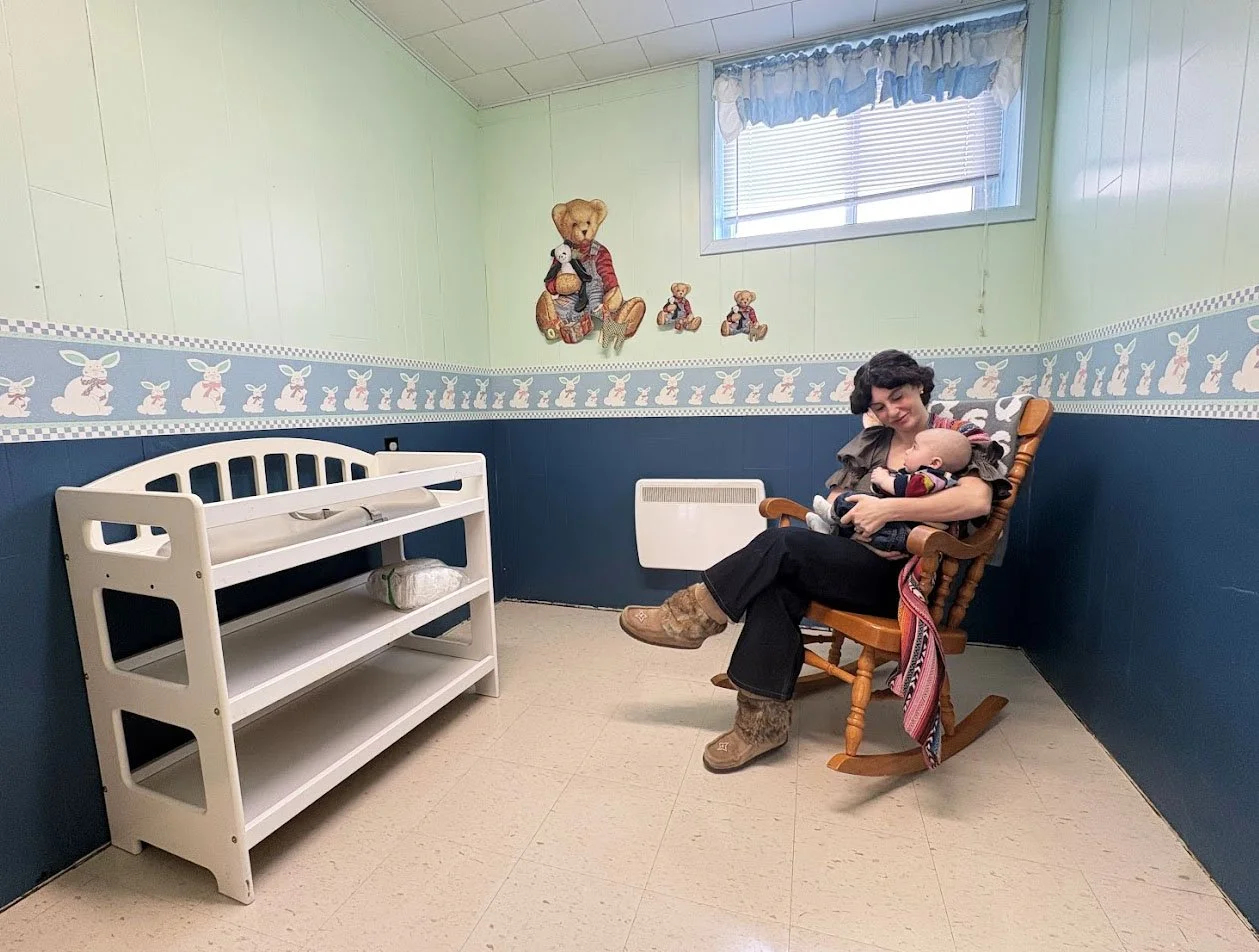A woman sitting in a rocking chair in a nursery, breastfeeding a baby, with bunny and teddy bear decorations on the wall and a window with blue curtains.