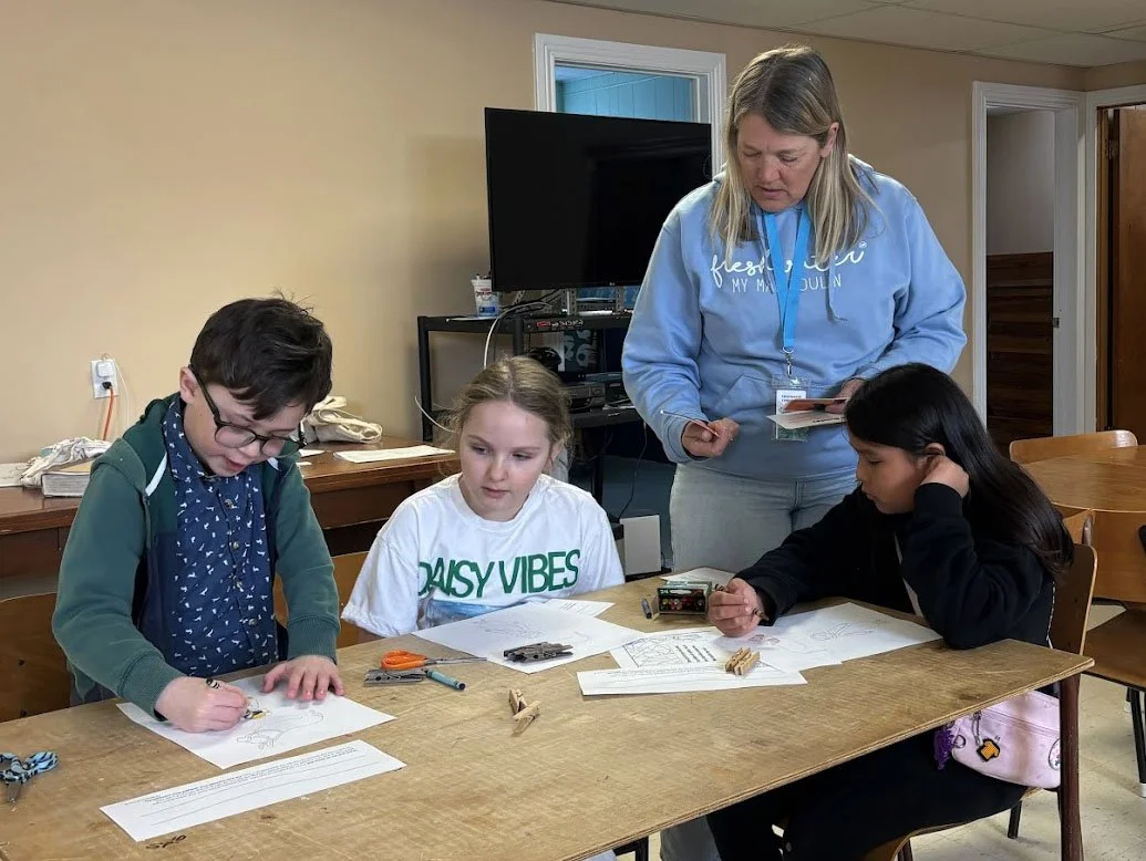 Three children sitting at a table with an adult woman standing behind them, engaged in a workshop or activity, with papers, clips, and small objects on the table.