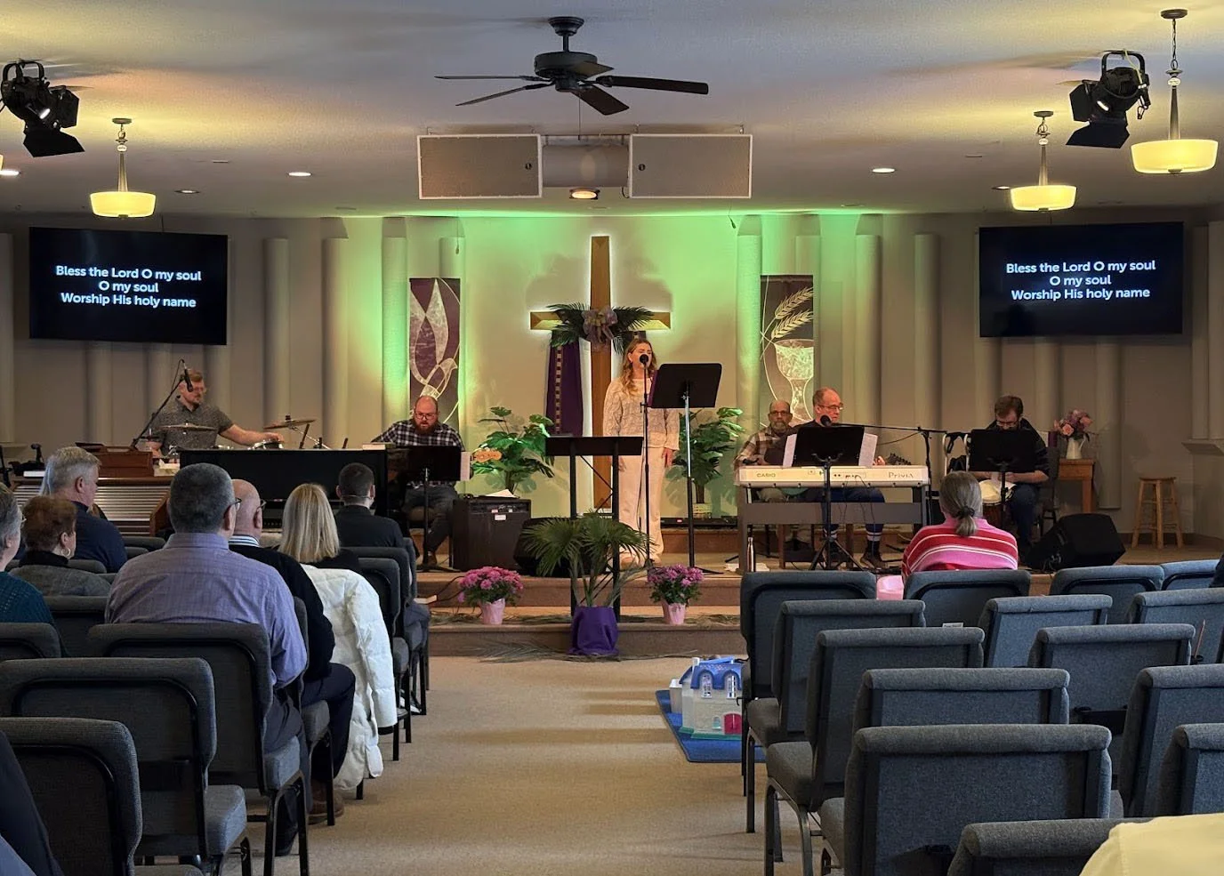 A church service or worship session taking place inside a church with a cross and religious decorations on the wall behind the stage. The stage has musicians, including a woman singing at a microphone, and screens on either side displaying song lyrics. The congregation is seated in rows of chairs, attentively listening.