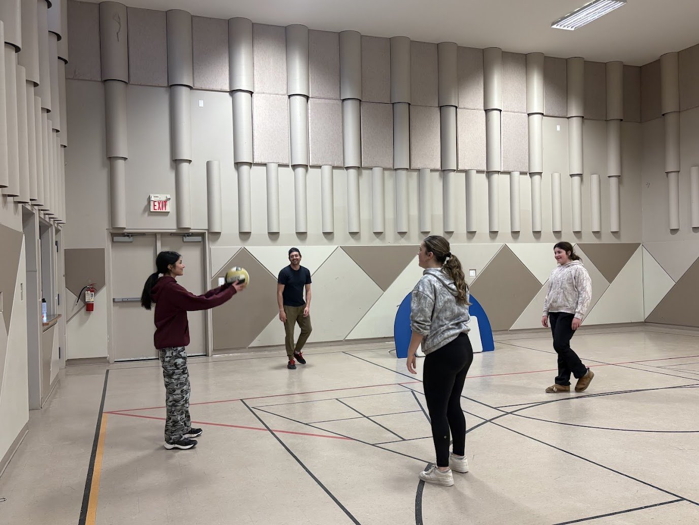 Four people playing volleyball indoors on a gymnasium court, with four players standing around, one of them holding a volleyball, and a blue goalpost in the background.