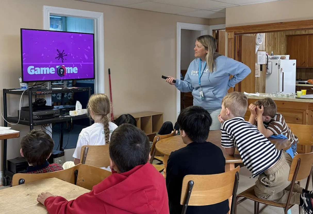 A woman standing in front of a group of children seated at tables in a room, with a TV screen displaying a game show called 'GameTime' in the background.