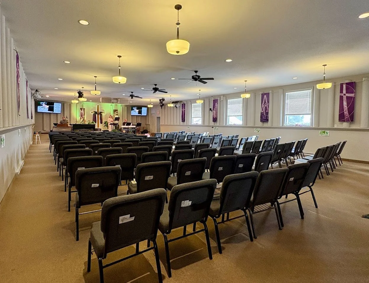 An empty church sanctuary with rows of black chairs facing a stage with musical instruments and screens. Purple banners hang on the walls, and windows allow natural light to enter.