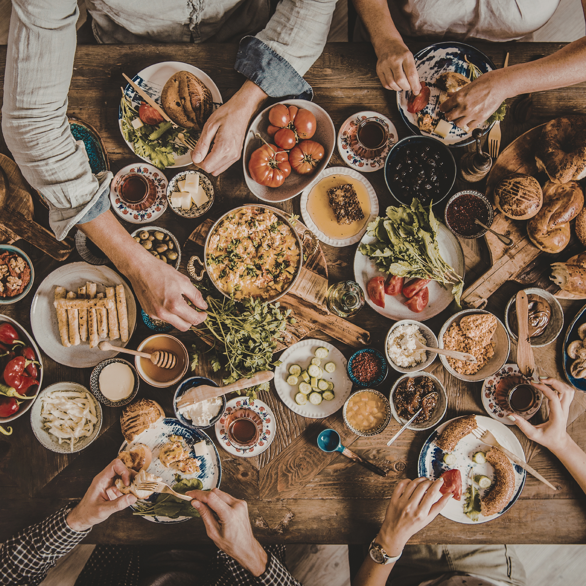 People sharing a large wooden table filled with various dishes, fruits, salads, bread, and drinks during a meal.