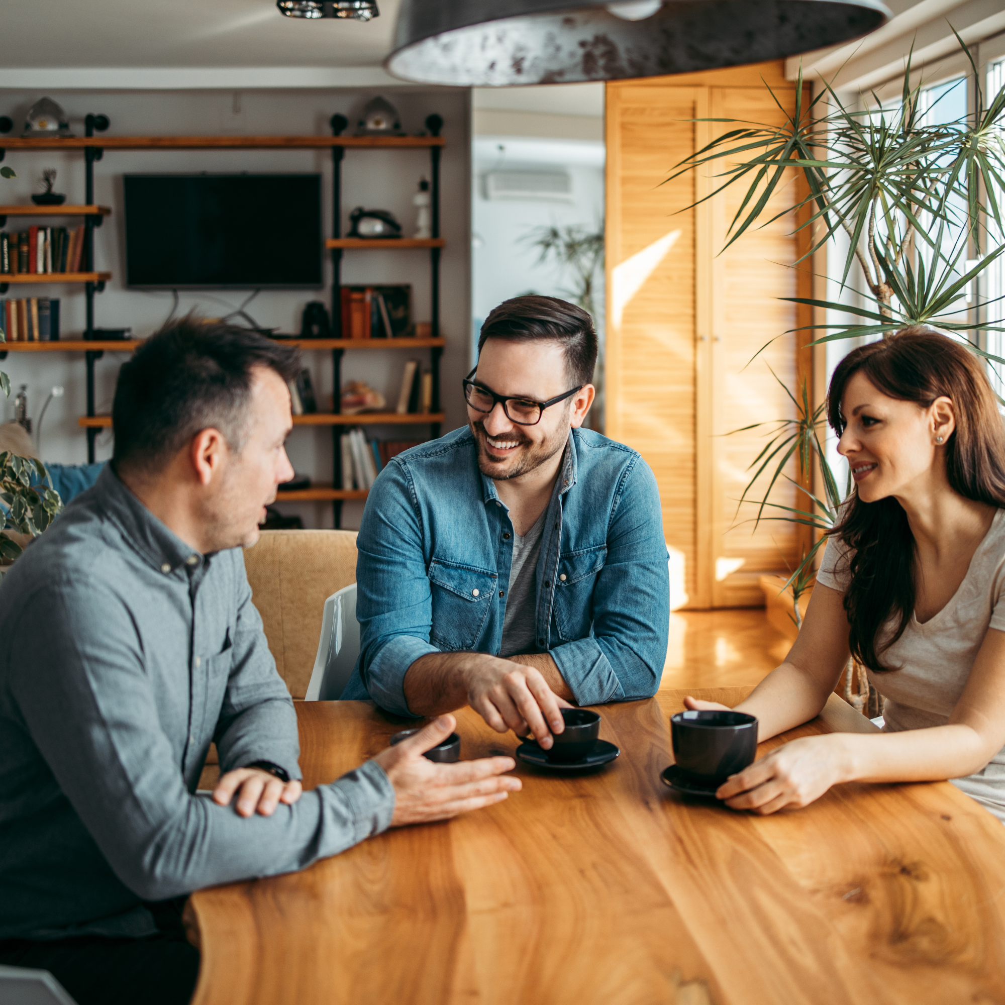 Three people sitting at a wooden table, smiling and talking, each with a black coffee mug. The setting is a cozy living room with bookshelves, plants, and a flat-screen TV.