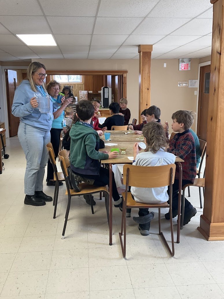 Children sitting around a table eating with adults standing nearby in a room with wooden paneling and an exit sign.