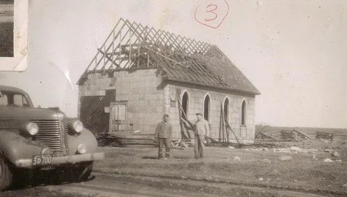 Black and white photo of a partially constructed brick building with a wooden roof frame, two men standing in front, and a vintage car parked nearby.