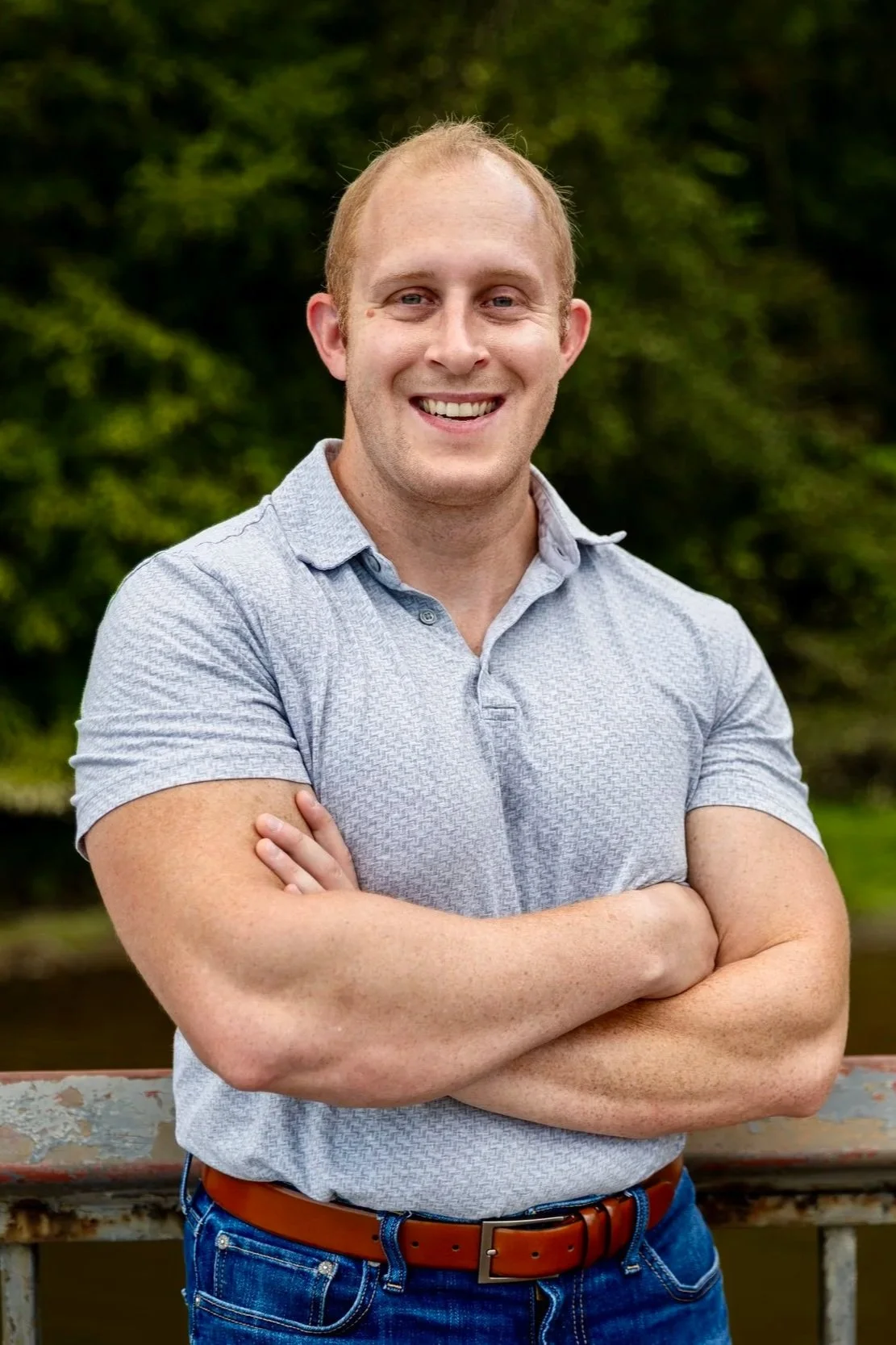 A smiling man with short light hair standing outdoors with his arms crossed, wearing a light gray polo shirt and blue jeans.