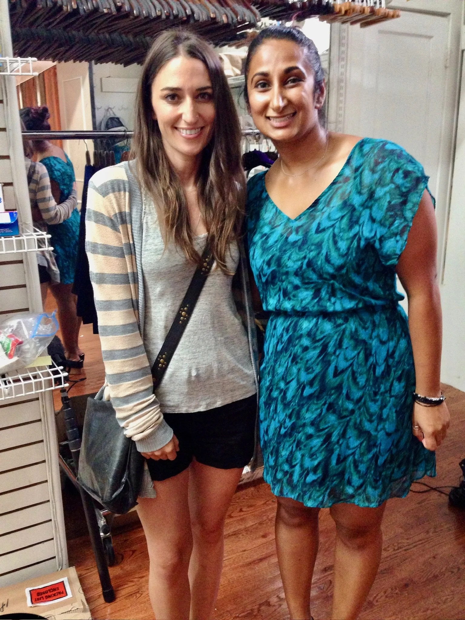 Two women standing close together and smiling for the camera in a retail store or clothing shop, with clothing racks and a person in the background. The Pearl Edge founder Parul "Pearl" Chokshi with Sara Bareilles shopping.