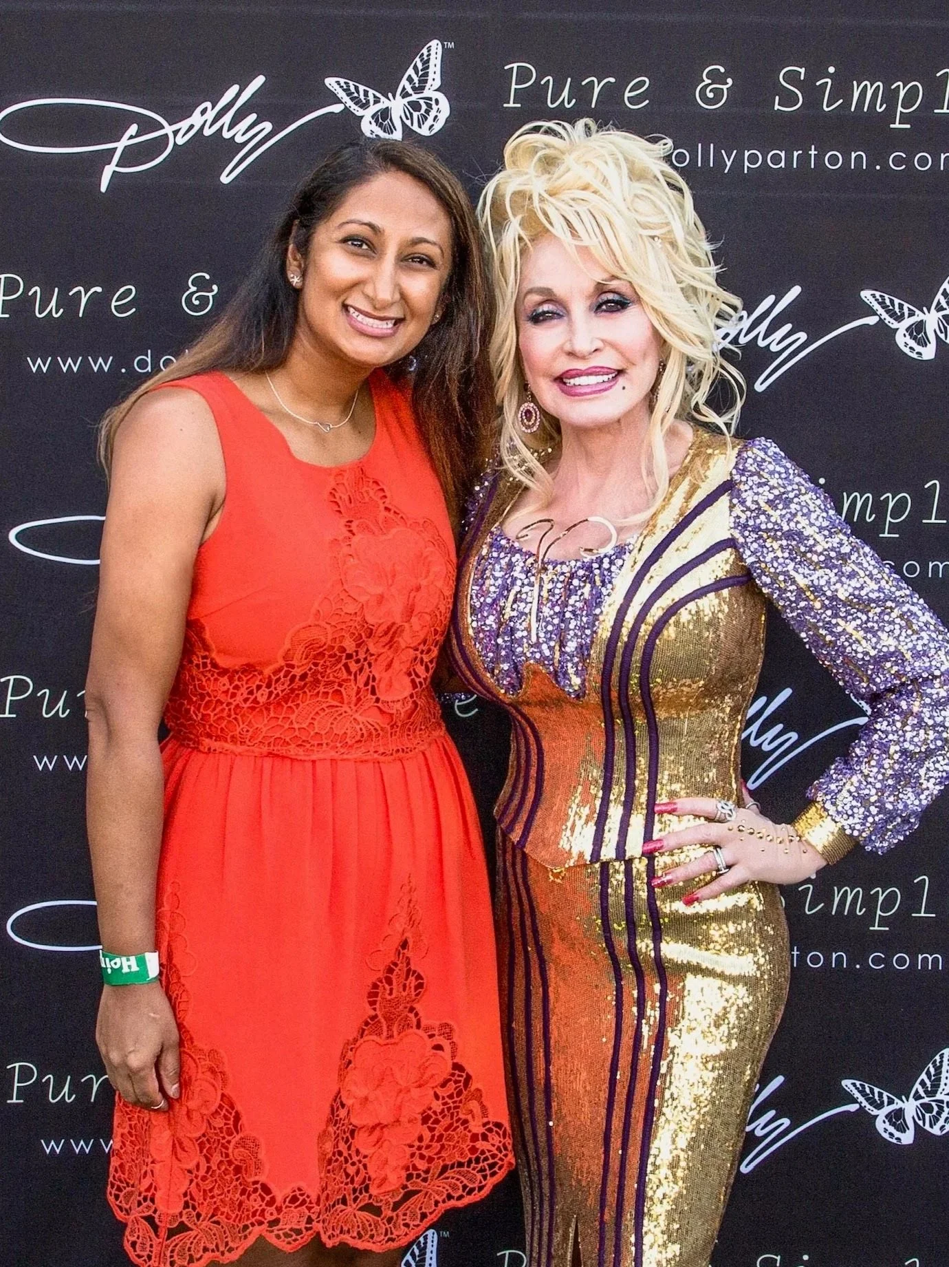 The Pearl Edge founder Parul "Pearl" Chokshi standing with Dolly Parton; one in a red dress with lace details and the other in a gold and purple sequined dress, posing at a promotional event with a black backdrop featuring butterfly graphics.