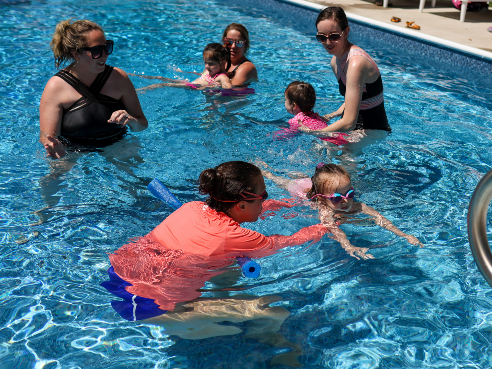 A group of women and children swimming and playing in a swimming pool on a sunny day.