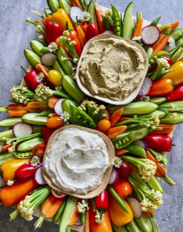 Assorted fresh vegetables surrounding two bowls of hummus, one beige and one white, on a large wooden platter.