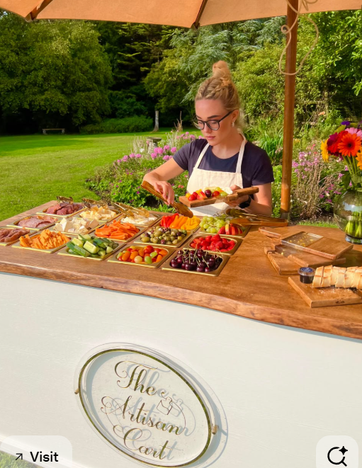 A woman serving herself at an outdoor fruit and vegetable buffet table, with a variety of fresh produce including grapes, cherry tomatoes, cut vegetables, and sliced meats, under a large umbrella with greenery and flowers in the background.