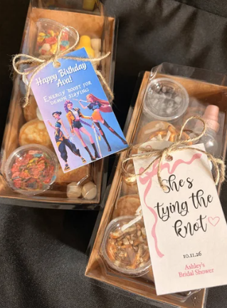 Two gift boxes filled with cookies and small candies, decorated with birthday tags. One tag says 'Happy Birthday Aunt' with a superhero image, and the other says 'She's tying the knot' with pink and heart decorations.