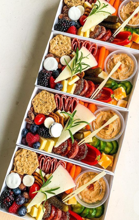 Four white trays filled with assorted foods including cheeses, crackers, grapes, berries, meats, vegetables, and small containers of dip or hummus.