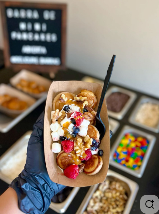 Cup of mini pancakes topped with syrup, whipped cream, mixed berries, marshmallows, and nuts, held in a hand wearing a black glove with a spoon; spread of toppings in bowls on a table in the background.