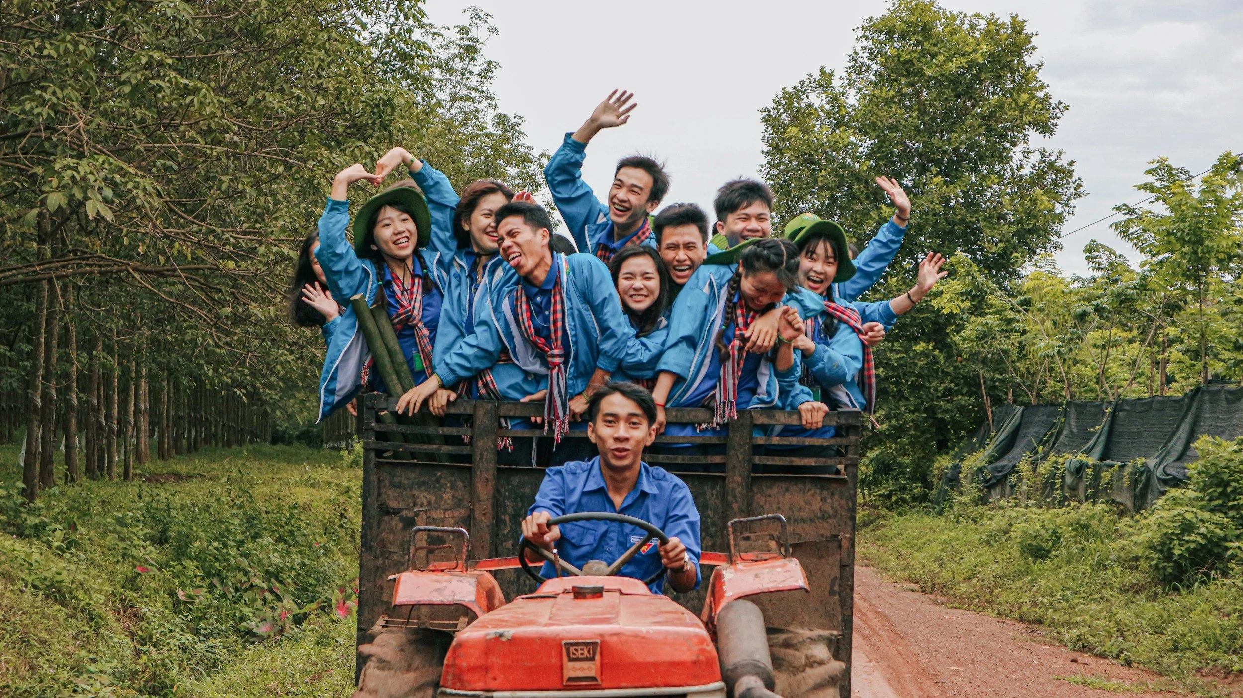Group of young people wearing blue uniforms and red neckerchiefs riding on a tractor through a lush, green farm, smiling and enjoying the ride.