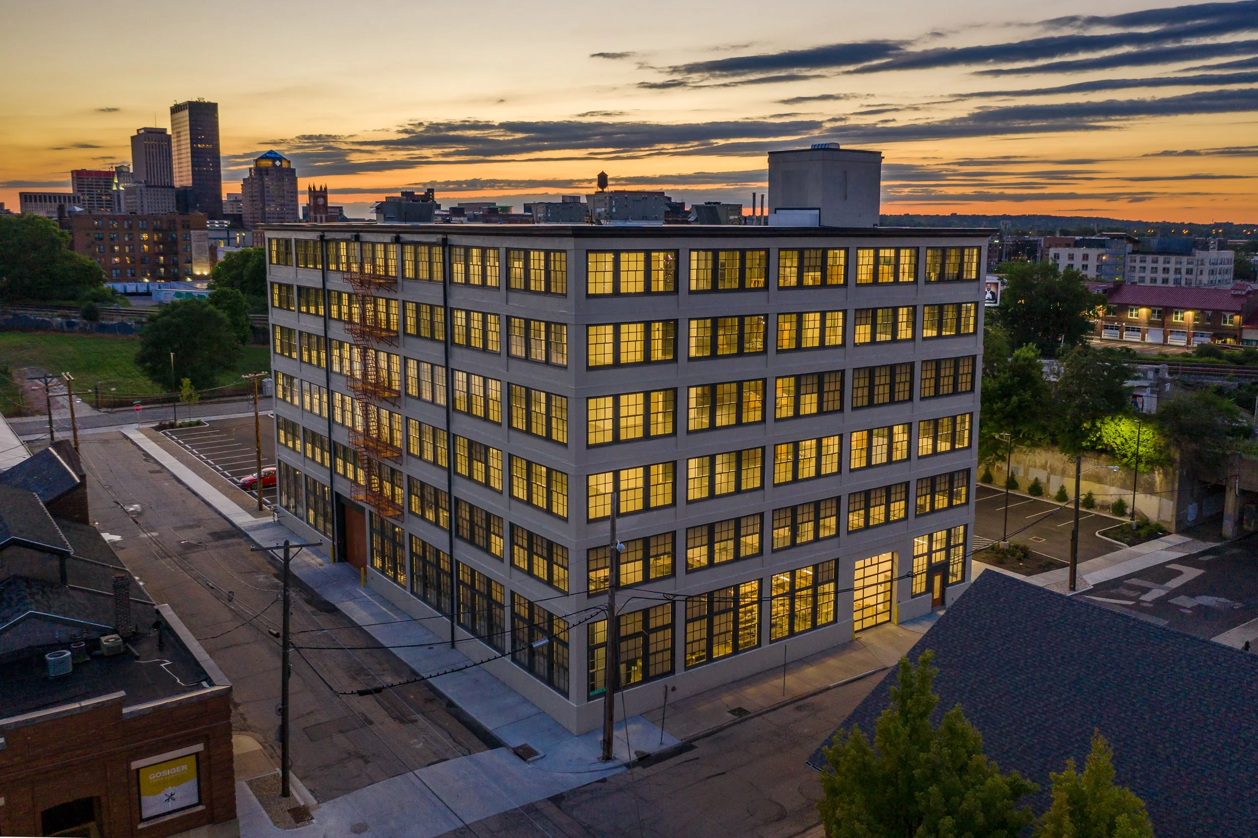 The Dayton Motor Car Building at dusk, a five-story historic concrete industrial building with rows of original multi-pane windows glowing warmly against a pink and blue sunset sky, situated on a corner lot in downtown Dayton, Ohio.