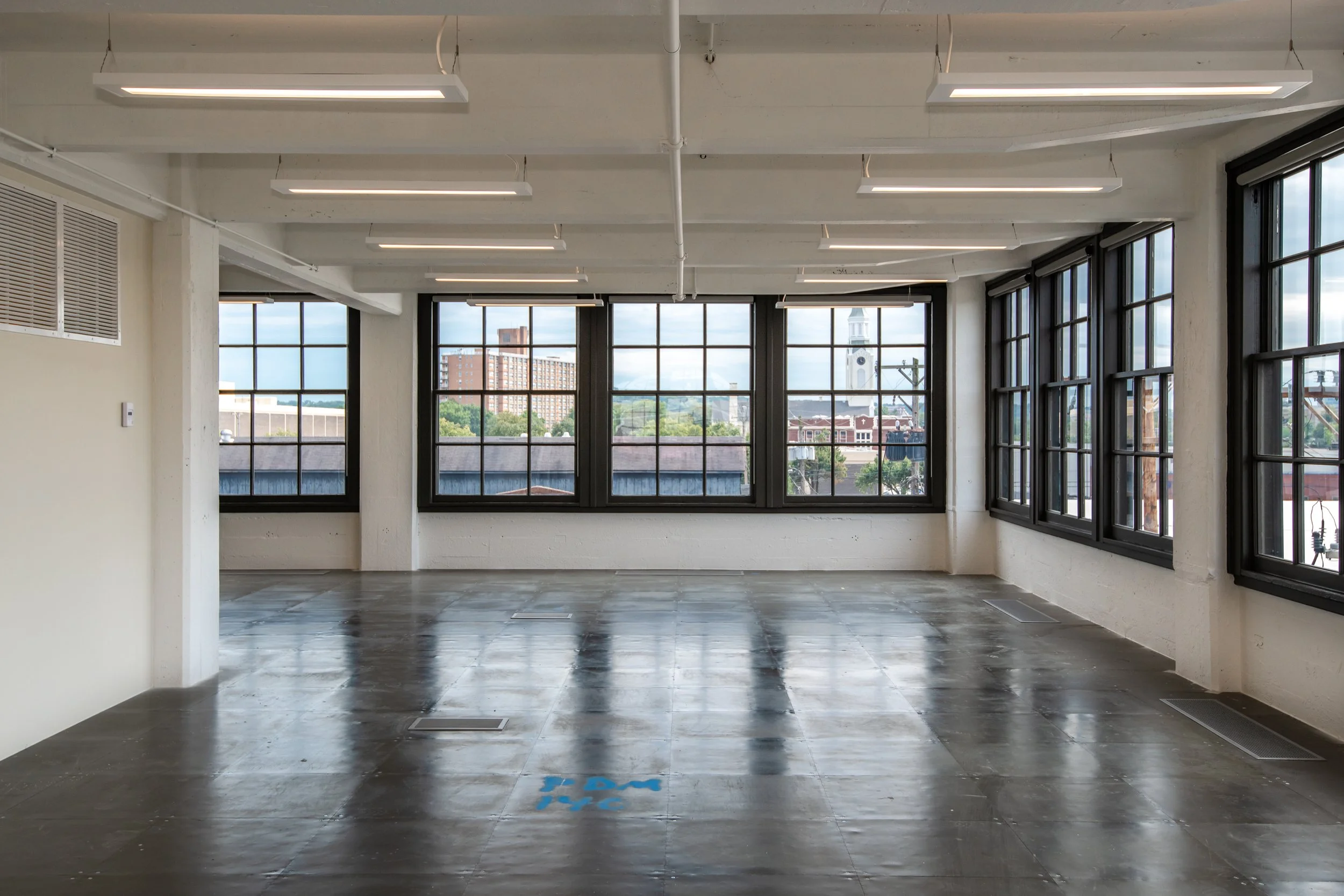 Empty room with large windows revealing cityscape and a church steeple, ceiling lights, and a concrete floor with the letters 'EPA' painted on it.