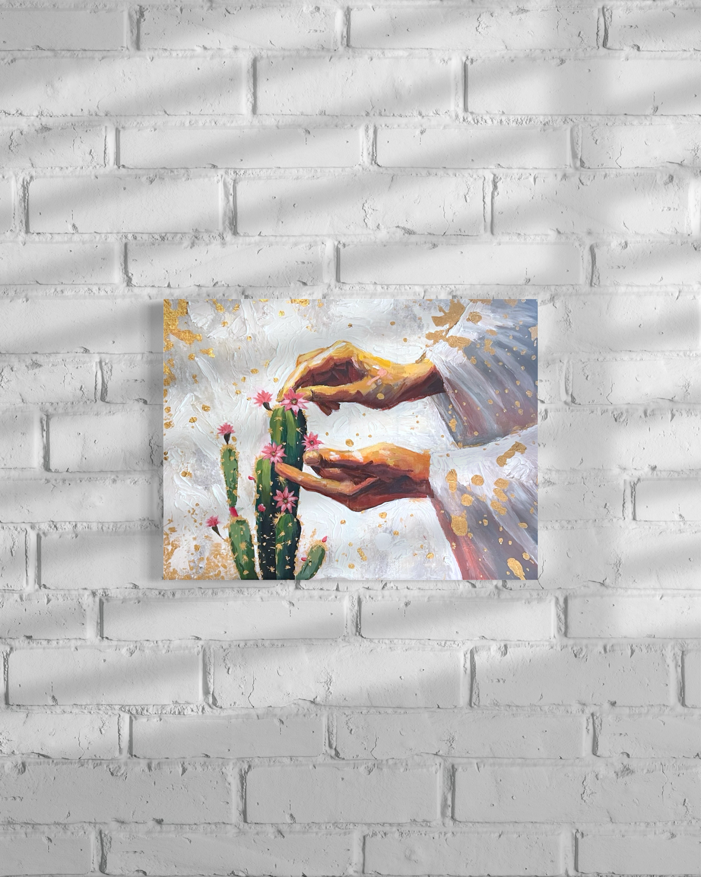 A painting of two hands forming a frame over a cactus with pink flowers, with gold splashes in the background.