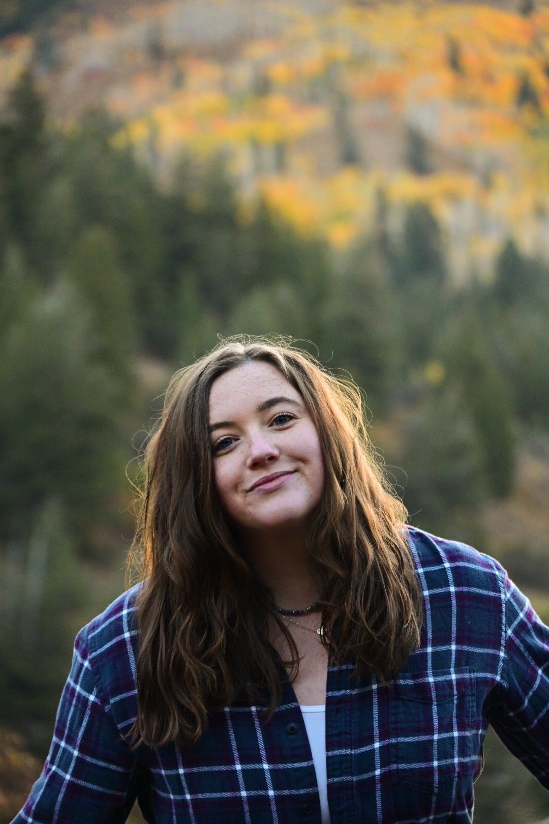 A young woman with long, wavy brown hair wearing a blue and red plaid shirt outdoors in a wooded area during autumn.