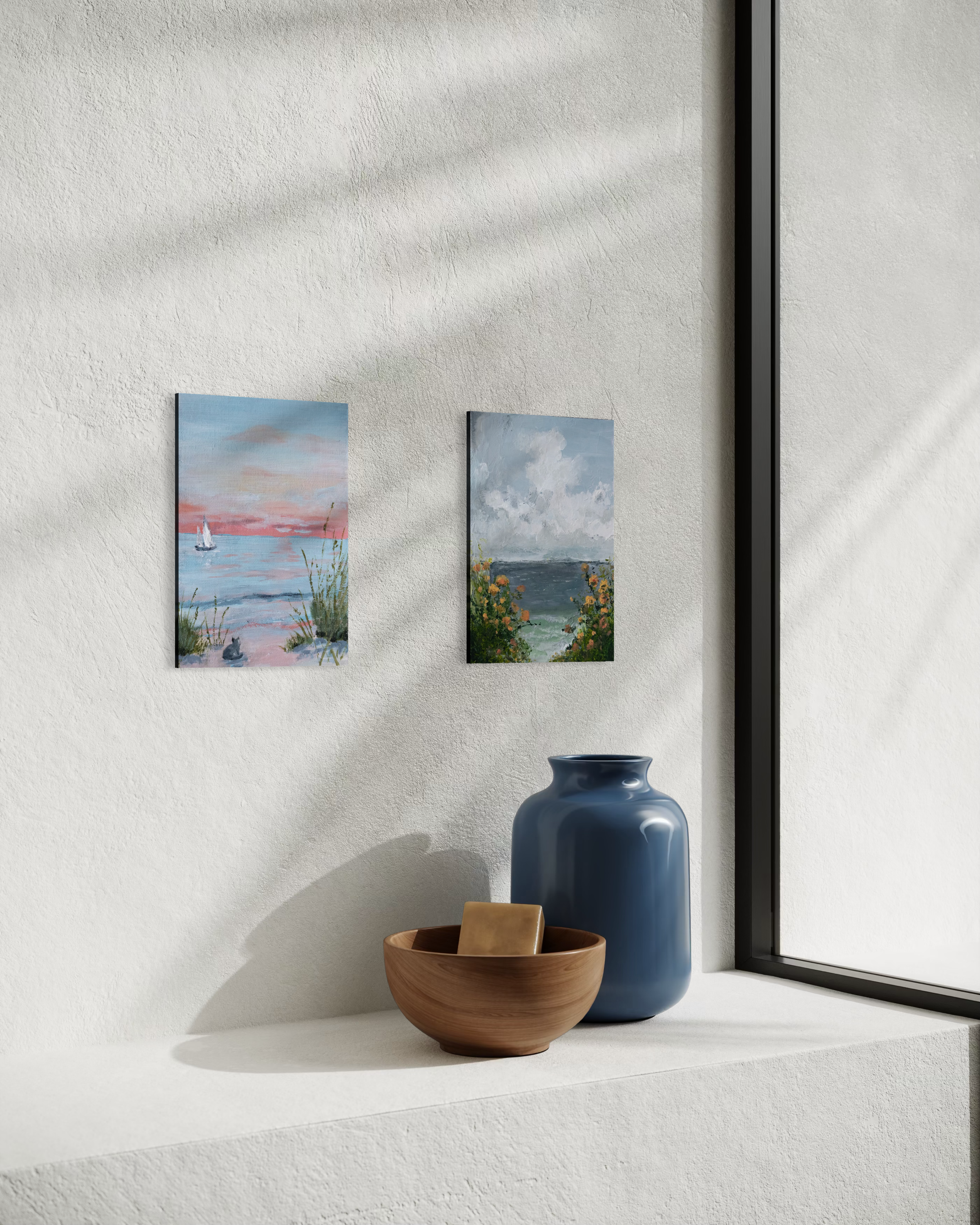 Interior with two small coastal landscape paintings on a white textured wall, a large blue vase, and a wooden bowl with a square object inside, next to a window.