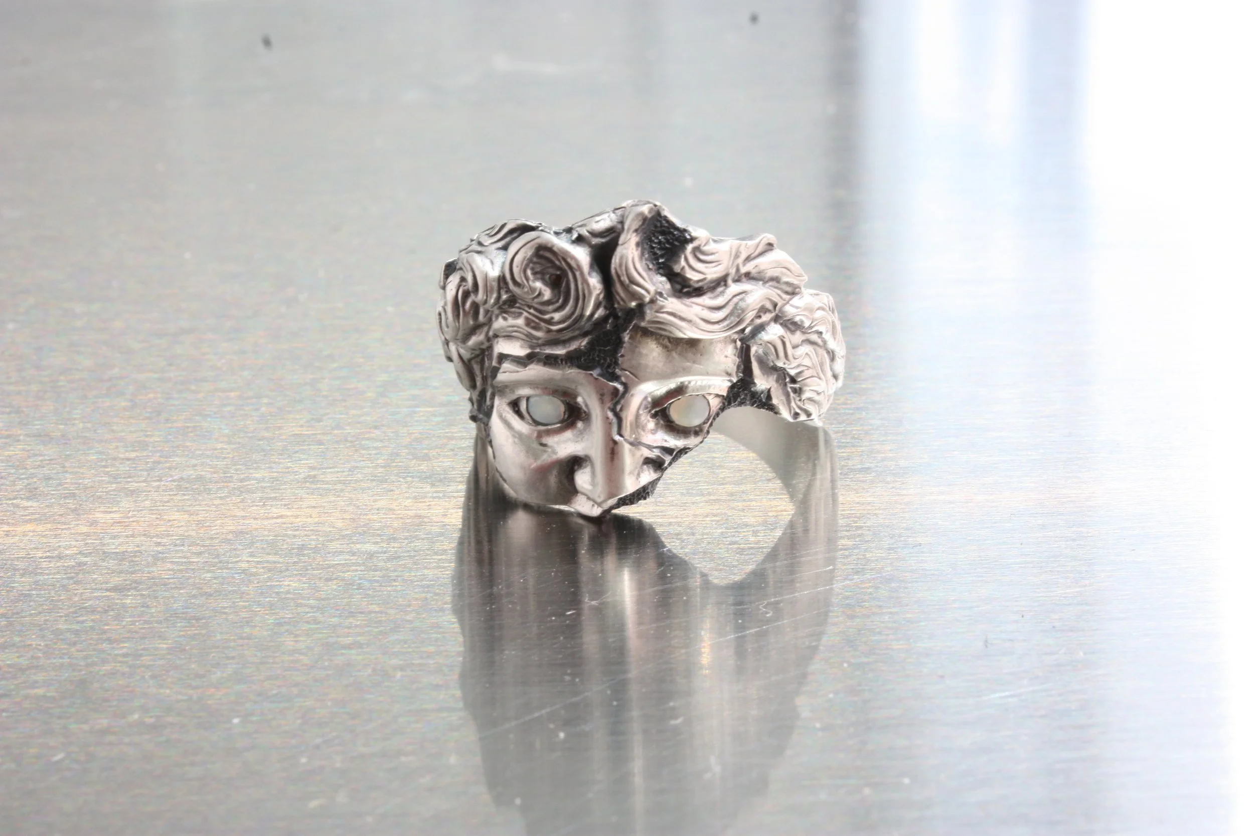 Anello con testa di Apollo in argento, con dettagli articolati opali cabochon incastonati negli occhi e riflessi metallici, posizionato su una superficie liscia e riflettente.