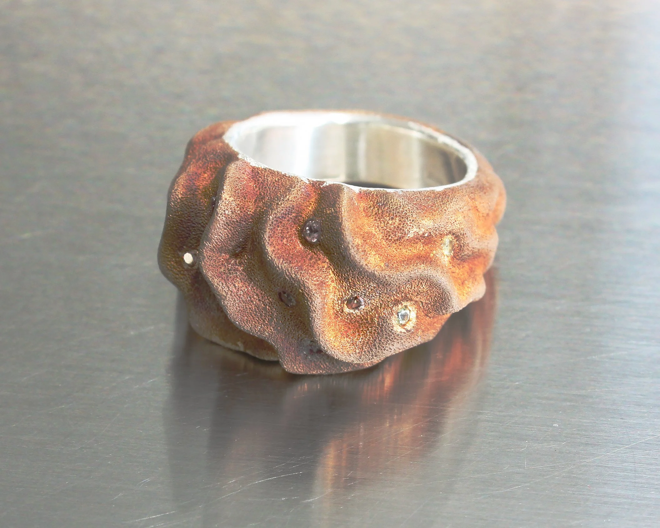 Anello scultoreo pezzo unico ELL Milano in bronzo e argento con zaffiri multicolore incastonati, posizionato su fondo inox metallico.
