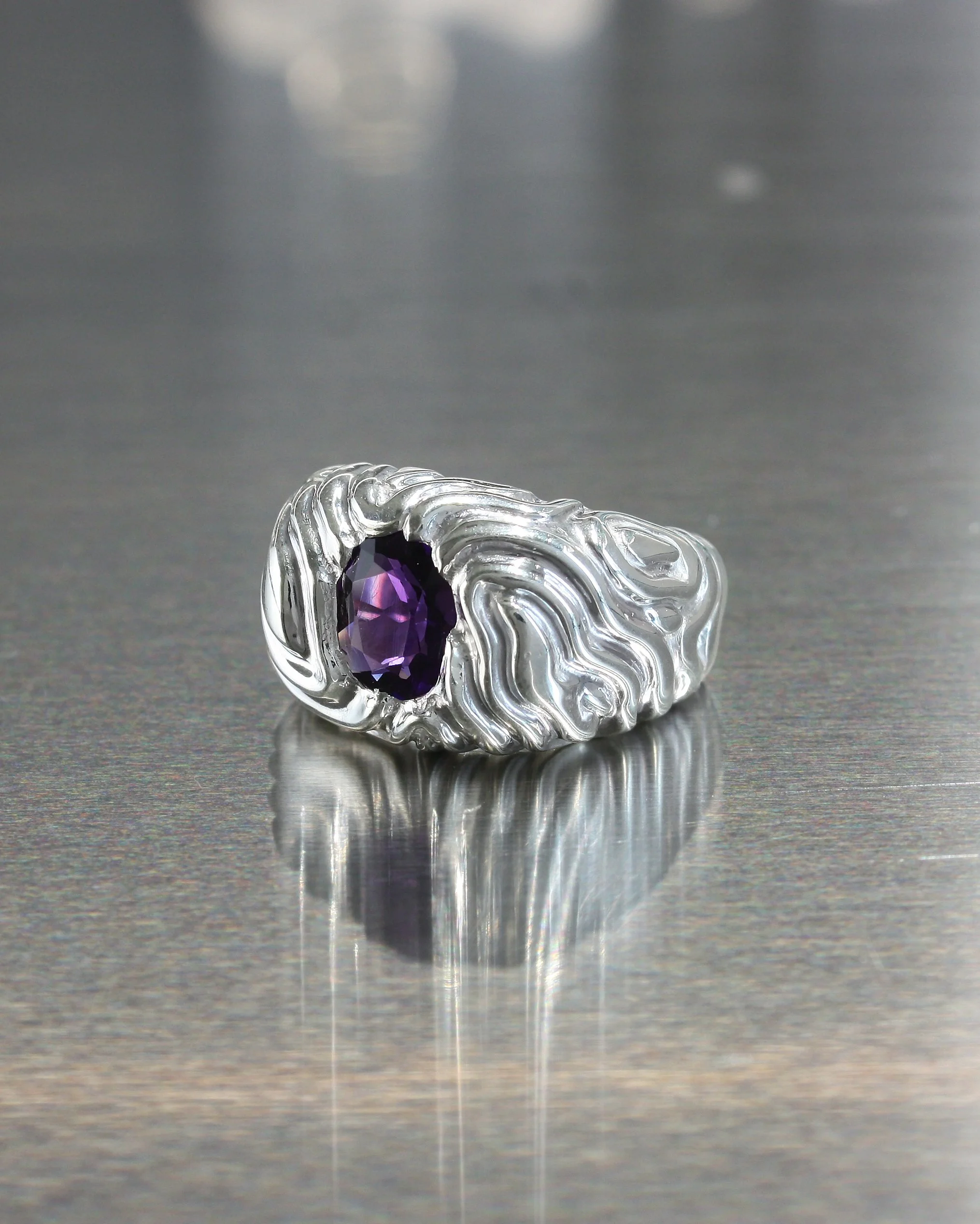 Anello scultoreo in argento lucido ELL Milano dalla superficie ondulata stile topografico trifimensionale con ametista taglio ovale. L'anello è posato su una superficie in inox.