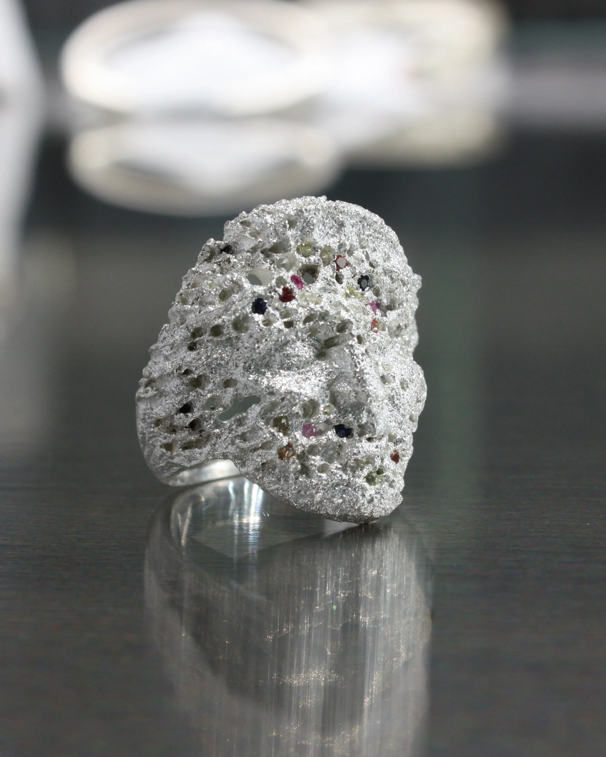 Anello ELL Milano in argento con texture porosa e irregolare incisa allover, decorato con piccoli zaffiri multicolore, posizionato sopra una superficie riflettente.