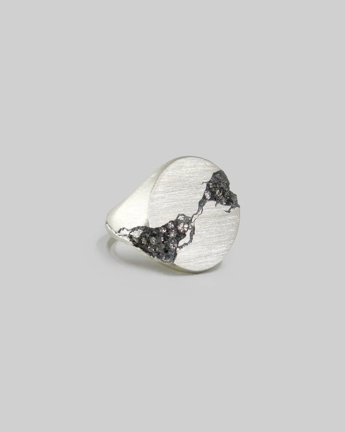 Anello a sigillo ovale in argento , con grosse fratture erose incise riempite da diamanti taglio brillante inacastonati, finitira brunita a contrasto con argento al naturale, fotografato su fondo neutro. 