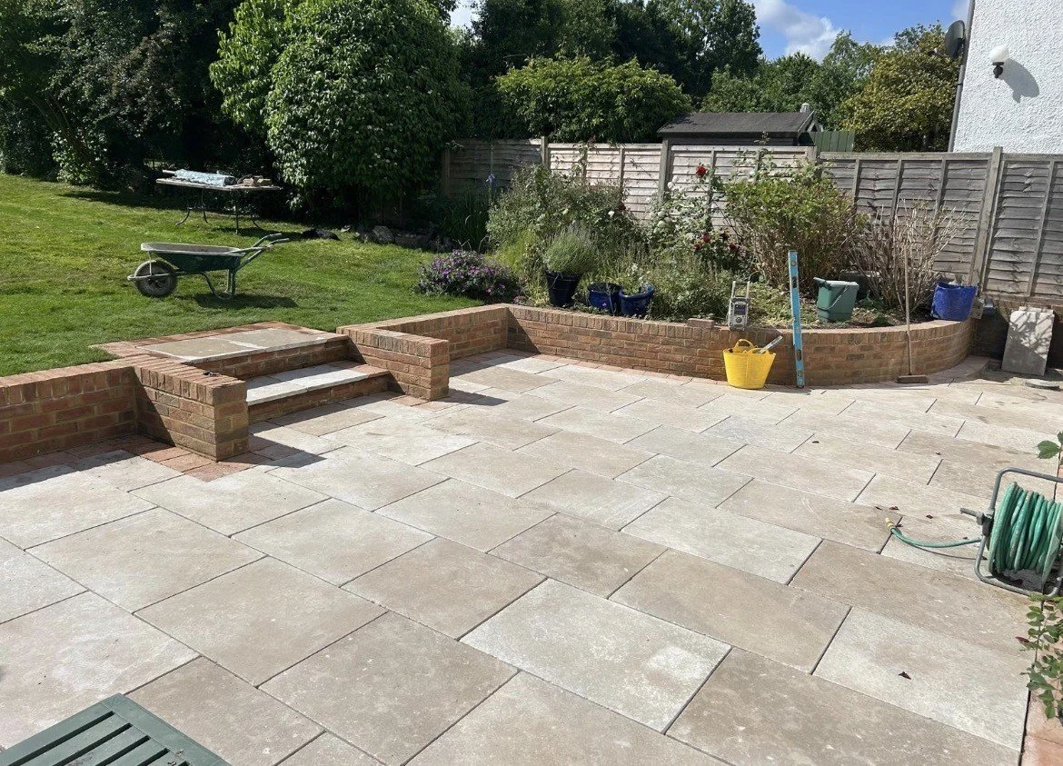 A backyard patio with newly laid paving stones, a brick raised garden bed with plants, gardening tools including a level, a yellow bucket, and a garden hose, and a grassy area with trees and shrubs in the background.