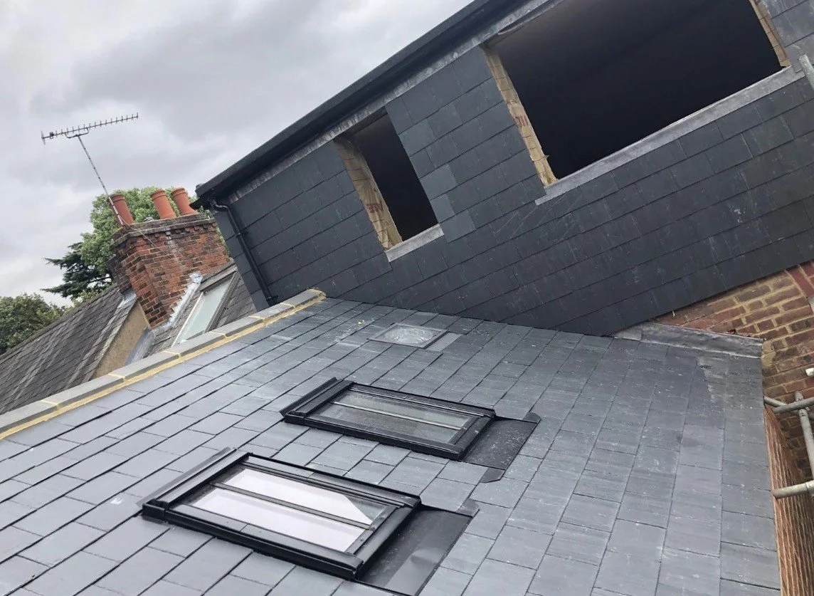 Roof with two skylights, dark slate tiles, and a brick chimney under a cloudy sky.