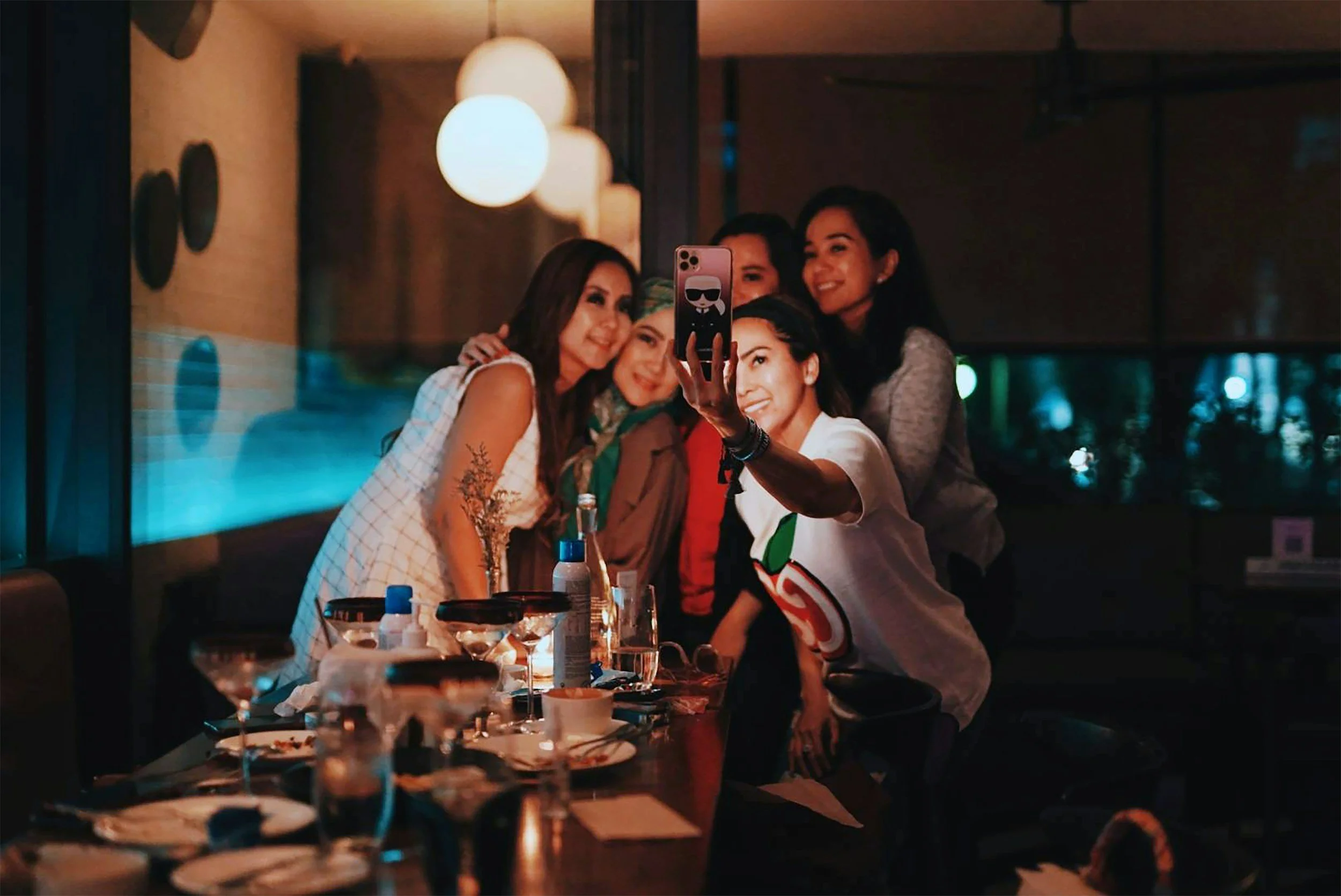 Group of five women taking a selfie at a restaurant or bar, with a table of drinks and food in front of them, in a dimly lit setting with hanging lights.