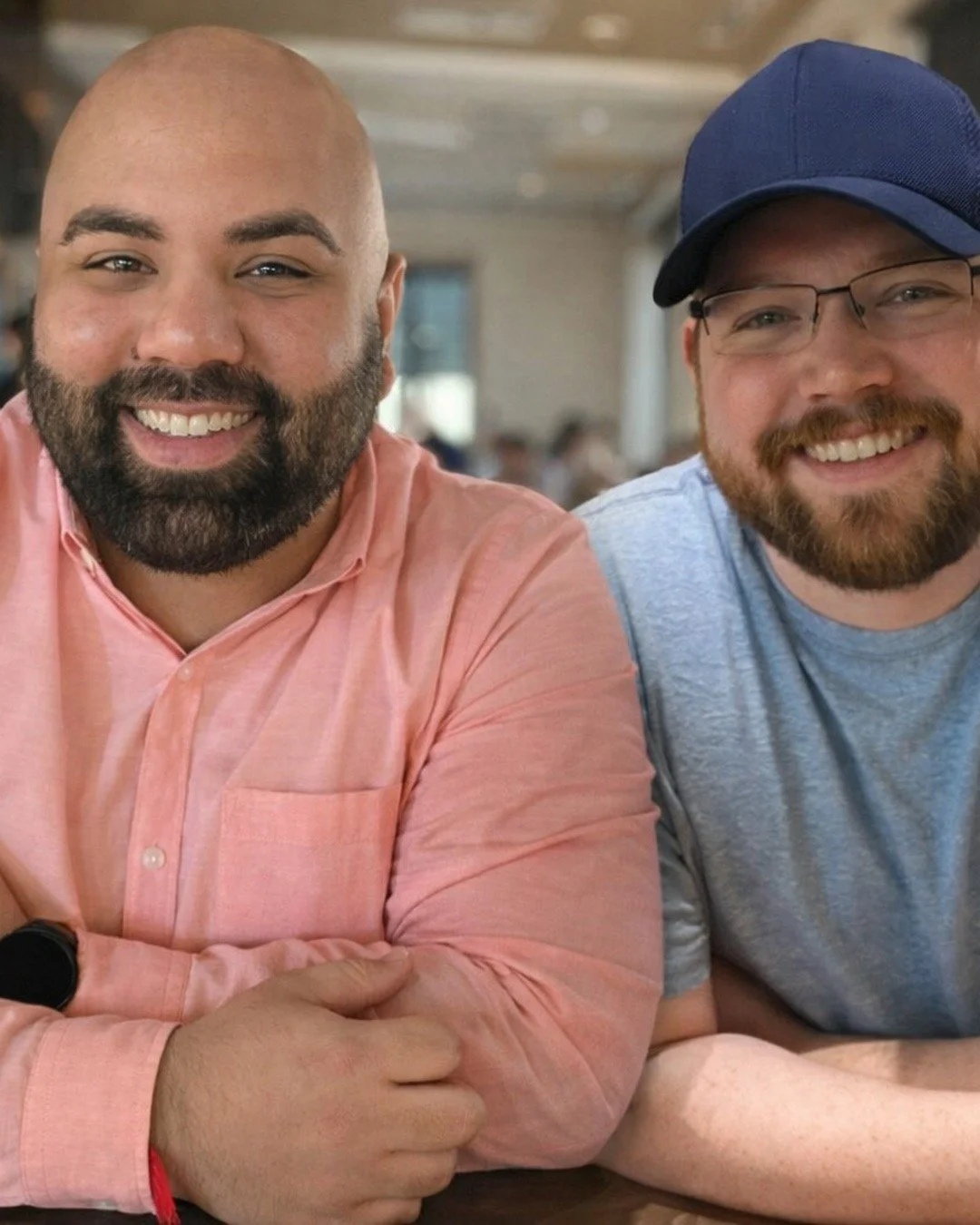 Two smiling men sitting at a table in a restaurant or cafe, with blurred background of other people and decor.