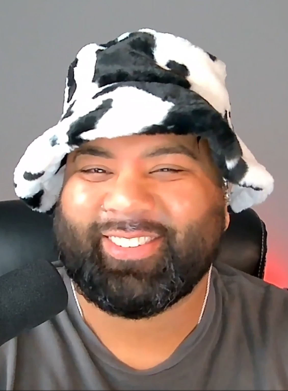 A man smiling with a beard, wearing a black and white cow print hat, sitting in front of a gray background and speaking into a microphone.