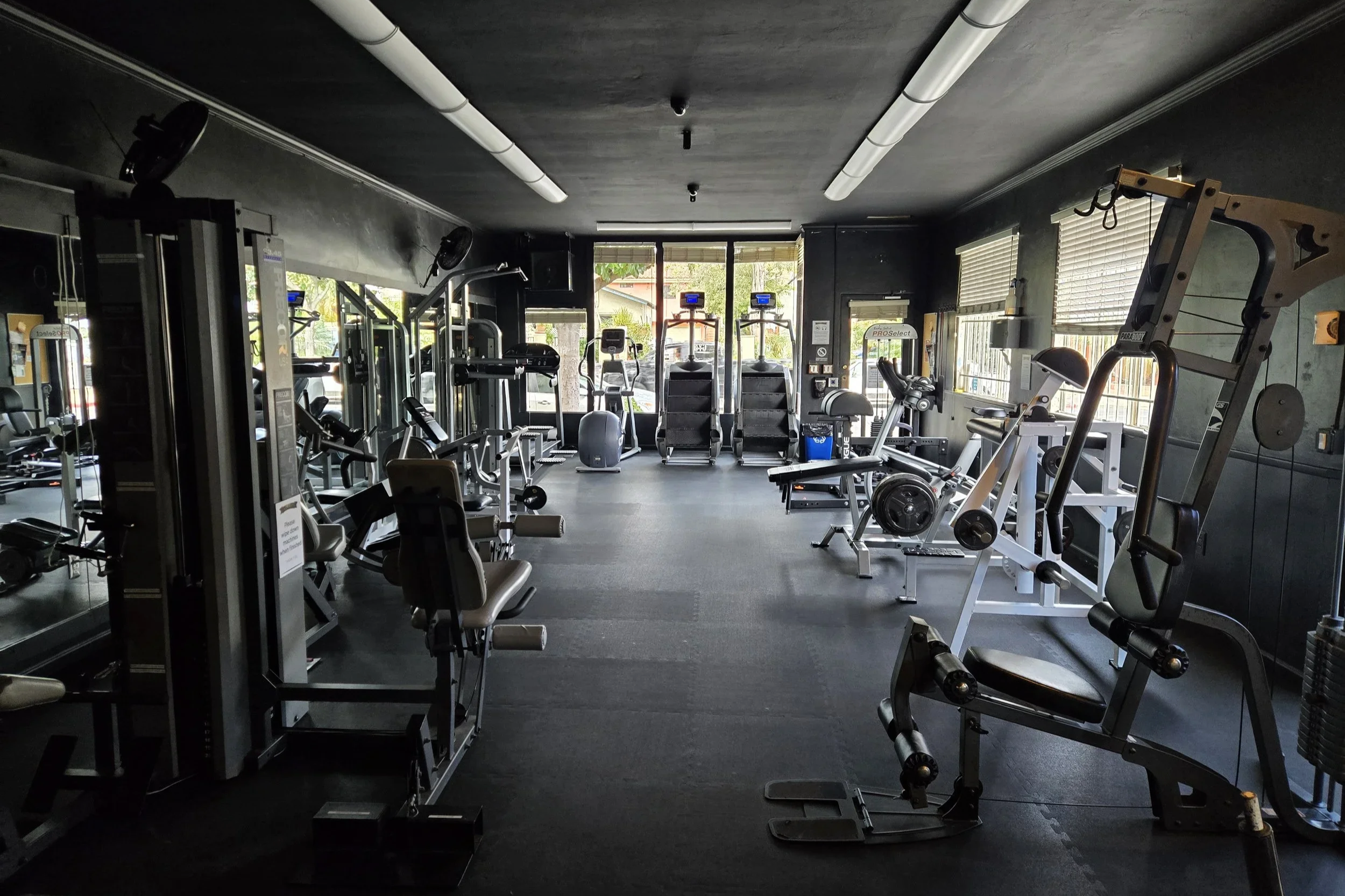 Interior of a fitness gym with various exercise equipment, including treadmills, ellipticals, weight machines, and dumbbells, with windows letting in natural light.